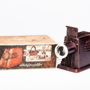 DIAR nejstarší vyráběná verze - červenohnědý bakelit.  Krabice s promítačkou a projekčním plátnem v černém podtisku, který měl vyvolávat pocit tmy v promítacím sále.